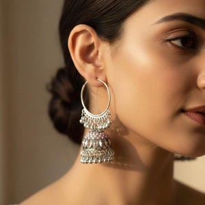 May include: Silver-toned hoop earrings with a dangling jhumka design. The earrings feature a large hoop, a decorative top section with small beads, and a bell-shaped bottom adorned with intricate patterns and more beads. The jhumka has a pink accent.