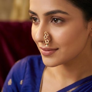 May include: A gold-toned nose ring with red and green gemstones and a small pearl. The jewelry is worn on the nose of a person wearing a blue top.
