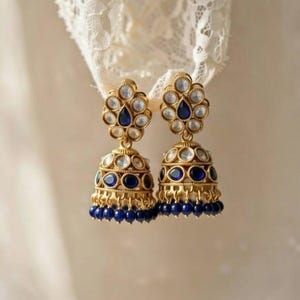 Może przedstawiać: Para złotych kolczyków z kwiatowym wzorem na górze i dzwonkowym kształtem na dole. Kolczyki mają niebieskie i przezroczyste kamienie szlachetne oraz małe niebieskie koraliki na dole. Wiszą na tle białej koronki.