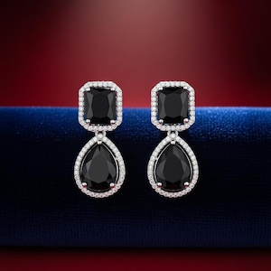 May include: A pair of elegant earrings featuring a square black gemstone at the top and a teardrop-shaped black gemstone below. The gemstones are surrounded by a halo of small, clear crystals, set in a silver-toned metal. The background is a gradient of red and blue.