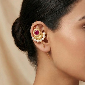 May include: A gold-toned ear cuff featuring a central ruby-colored stone surrounded by small pearls. The design includes intricate detailing and a long, straight bar that extends down the ear.
