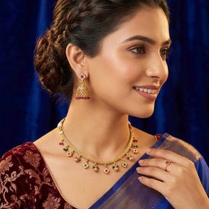 May include: A woman wearing a gold necklace, earrings, and ring set with red, green, and white gemstones. The necklace and earrings have a floral design. The woman is wearing a maroon velvet top and a blue sari.