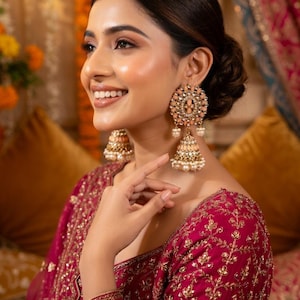 Puede incluir: Una mujer lleva pendientes grandes y ornamentados con un diseño melocotón y dorado. Los pendientes tienen una parte superior circular con un diseño floral y perlas colgantes. Lleva un top bordado burdeos.