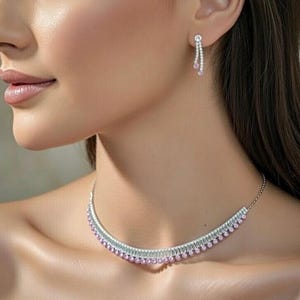 May include: A silver necklace and matching earrings set. The necklace is a choker style with a row of pink gemstones and a row of clear stones. The earrings have a similar design, with a single clear stone at the top and a dangling row of pink gemstones.