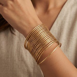 Può includere: Un set di numerosi bracciali rigidi circolari e sottili in metallo color oro indossati insieme al polso. I bracciali presentano una finitura liscia e lucida con un profilo uniforme e sottile, creando un aspetto sovrapposto sull'avambraccio.