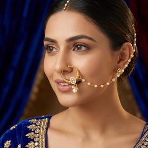 May include: A gold-toned nose ring featuring a circular design with white stone accents and a dangling pearl-like bead. A delicate gold chain adorned with small white beads connects the nose ring to a matching earring piece worn on the ear.