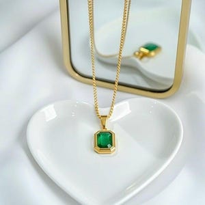 May include: A gold-colored necklace with a rectangular emerald-green pendant. The pendant is set in a gold frame and hangs from a delicate gold chain. The necklace is displayed on a white heart-shaped dish.