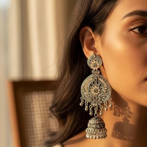 May include: Silver-toned, ornate earrings with a floral design. The earrings feature a large, detailed pendant with smaller dangling elements and a bell-shaped bottom. The design includes intricate patterns and small, round accents, creating a vintage aesthetic.