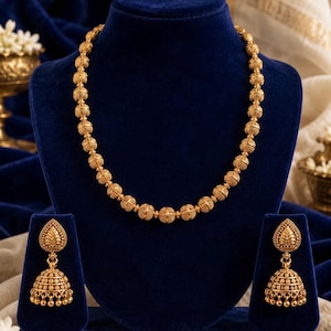 May include: A gold-toned jewelry set consisting of a necklace and two matching earrings. The necklace features a series of round, textured beads. Each earring includes a teardrop-shaped top piece connected to a bell-shaped jhumka with small beads.