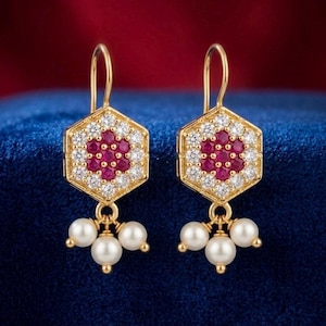 May include: A pair of gold-toned earrings. Each earring features a hexagonal top with a central cluster of red gemstones surrounded by white gemstones. Three dangling pearls hang from the bottom of each earring.