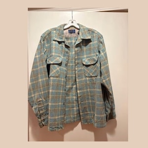 May include: A long-sleeve button-up shirt featuring a plaid pattern in shades of light blue and tan. The shirt includes a pointed collar, two chest pockets with buttoned flaps, and a front button closure. A dark blue brand label is visible at the neck.