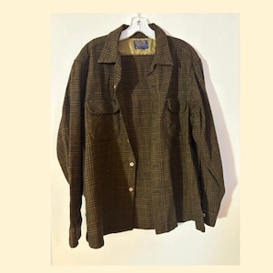 May include: A long-sleeved button-down shirt in a dark olive green and black plaid pattern. The shirt features a pointed collar, two chest pockets with buttoned flaps, and a light tan interior lining visible at the neck with a Pendleton brand label.