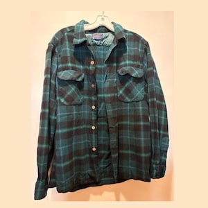 Peut inclure: Une chemise à manches longues boutonnée avec un motif à carreaux vert foncé et noir. La chemise présente un col pointu, deux poches de poitrine à rabat boutonné et une rangée verticale centrale de boutons. Elle est sur un cintre blanc.