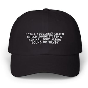 Op de afbeelding: Zwarte baseballcap met witte tekst: "I STILL REGULARLY LISTEN TO LCD SOUNDSYSTEM'S SEMINAL 2007 ALBUM 'SOUND OF SILVER'". De cap heeft een gebogen klep en een knoop aan de bovenkant.