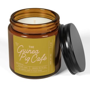 May include: A brown glass candle jar with a black lid. The label reads "The Guinea Pig Cafe" with the text "Smells Like" and "With notes of emotional chaos, breaking the 4th wall, the lingering scent of stolen statues." The candle is 100% soy wax and is 4 oz.