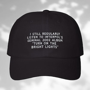 May include: Black baseball cap with white embroidered text that reads "I still regularly listen to Interpol's seminal 2002 album "Turn on the Bright Lights""