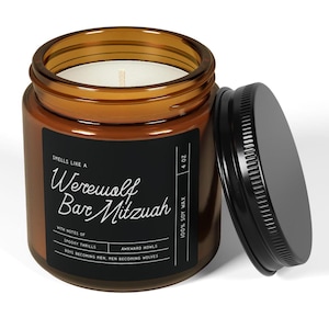 Puede incluir: Un tarro de vela de vidrio marrón con una etiqueta negra que dice "Smells like a Werewolf Bar Mitzvah" con notas de "Spooky Thrills" y "Awkward Howls". La vela es 100% cera de soja y pesa 4 oz.
