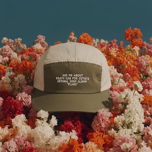 May include: A five-panel cap with a cream-colored top and a dark olive green bill and front panel. The front panel features the text "ASK ME ABOUT DEATH CAB FOR CUTIE'S SEMINAL 2005 ALBUM 'PLANS'" in white. The cap is surrounded by colorful flowers.