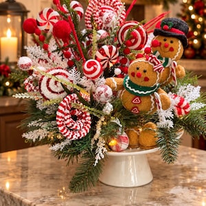 May include: A holiday centerpiece on a white pedestal featuring two gingerbread figures, one wearing a black hat, nestled among evergreen branches, red and white peppermint candy ornaments, and frosted white pine sprigs with red berries.