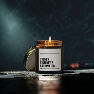 May include: A lit candle in a glass jar with a gold lid and a label that reads "SYDNEY SWEENEY'S BATHWATER". The candle is on a dark surface with a dark background. The jar is open, with the lid next to it.