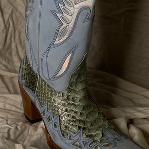 Può includere: Un paio di stivali da cowboy blu chiaro con un motivo a pelle di serpente verde sul gambale e sulla punta. Gli stivali hanno una punta a punta e un tacco alto.