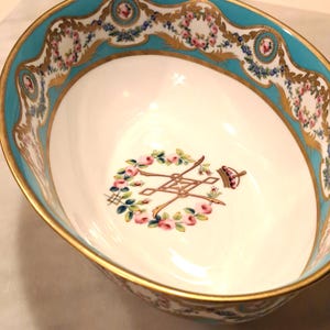 May include: Oval porcelain bowl with a white interior and a turquoise rim. The rim is decorated with gold filigree and floral patterns. The bowl's interior features a floral wreath design with gold accents and a crown.