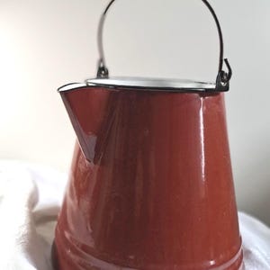 May include: A vintage, red-brown enamelware kettle with a black handle and spout. The kettle has a classic design with a rounded body and a curved handle. The spout is angled for easy pouring. The kettle is likely used for boiling water.