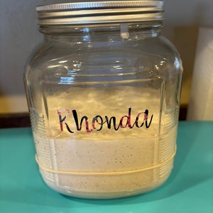 May include: A clear glass jar with a silver lid, containing a light-colored substance. The jar has the name "Rhonda" in a floral design. A rubber band encircles the jar. The jar is on a turquoise surface.