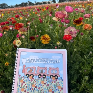 以下が含まれることがあります： 表紙に「Lili's Adventures」というタイトルが書かれたピンク色のスパイラルバインディングされた本。表紙には、ピンクの電車の中に3匹のクマが乗っているという漫画のイラストが描かれている。電車はカラフルな花畑の中を走っている。