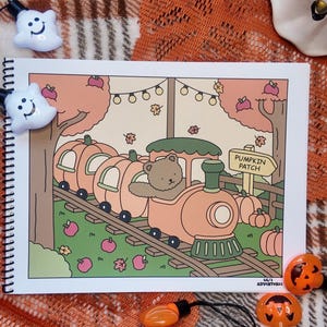 May include: A spiral-bound sketchbook featuring a cartoon illustration of a brown bear driving a pumpkin-shaped train through a pumpkin patch. The scene includes autumn trees, falling leaves, string lights, and a sign labeled PUMPKIN PATCH.