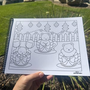 May include: A spiral-bound coloring book featuring line art illustrations of teddy bears in teacups, with bows and decorative elements. The book includes the text "ULI'S ADVENTURES" at the bottom right corner. The image is held outdoors.