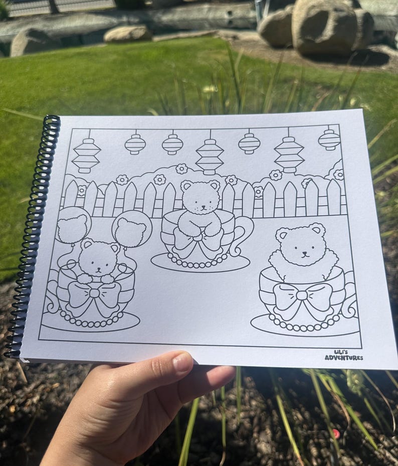 May include: A spiral-bound coloring book featuring line art illustrations of teddy bears in teacups, with bows and decorative elements. The book includes the text "ULI'S ADVENTURES" at the bottom right corner. The image is held outdoors.