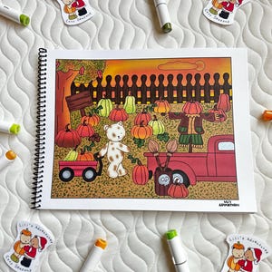 May include: A spiral-bound sketchbook featuring a colorful autumn illustration with pumpkins, a teddy bear, and a scarecrow. Scattered around are art markers and stickers with the text "Lili's Adventures Cozy Seasons."