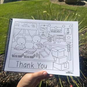 May include: A black and white coloring book with a spiral binding, held by a hand. The cover reads "Thank You" in large letters. Inside, there are illustrations of bears, boxes, and a desk with the text "Lil's Adventures".