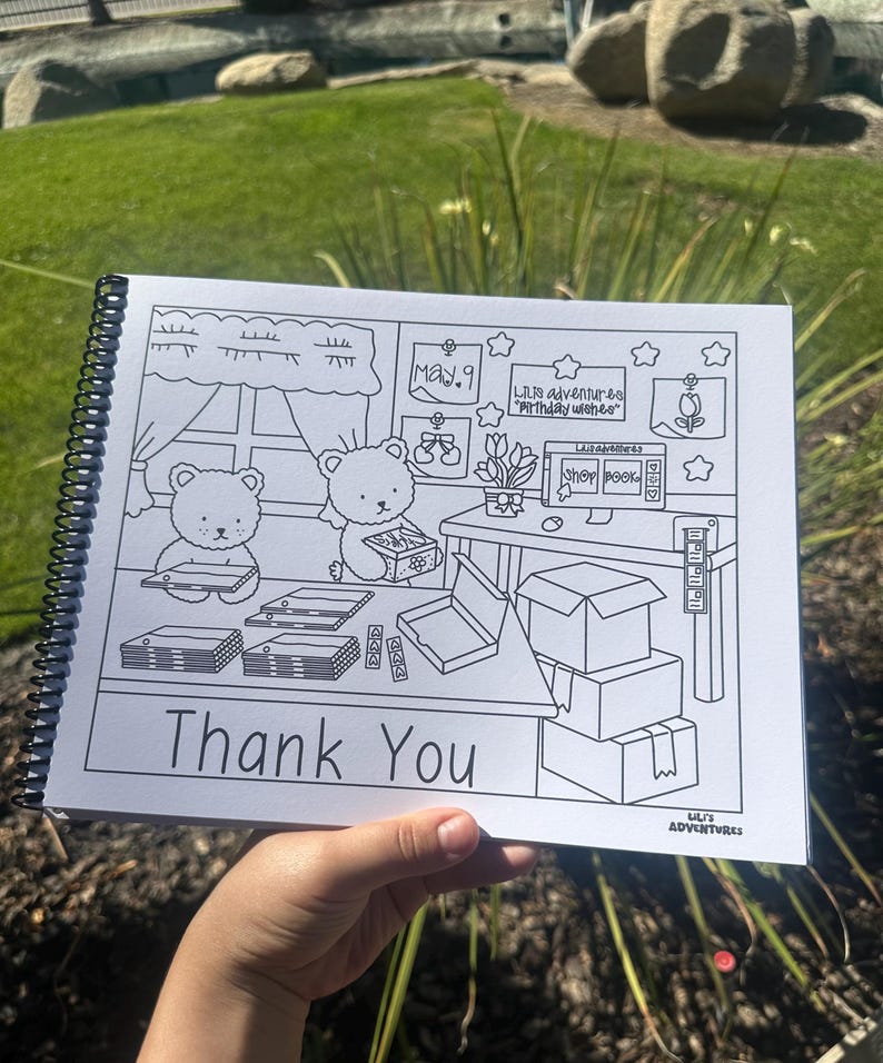 May include: A black and white coloring book with a spiral binding, held by a hand. The cover reads "Thank You" in large letters. Inside, there are illustrations of bears, boxes, and a desk with the text "Lil's Adventures".