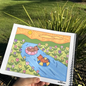 May include: A spiral-bound coloring book featuring a whimsical illustration of two brown bears floating on inner tubes in a blue river. The river flows through a field of colorful flowers under a yellow sun. The text "Lili's Adventures" is in the bottom right corner.