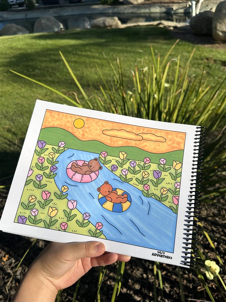 May include: A spiral-bound coloring book featuring a whimsical illustration of two brown bears floating on inner tubes in a blue river. The river flows through a field of colorful flowers under a yellow sun. The text "Lili's Adventures" is in the bottom right corner.