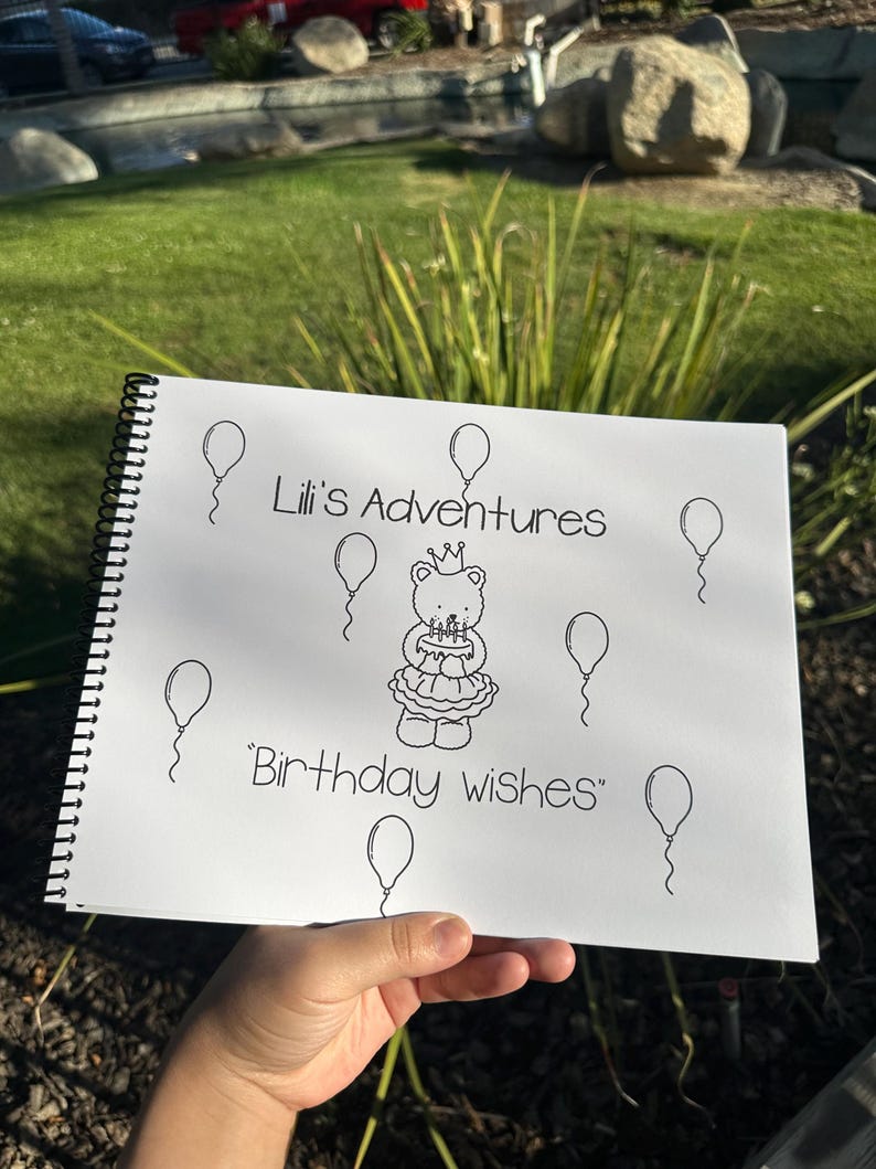 May include: A spiral-bound notebook with the text "Lili's Adventures" and "Birthday wishes" in black ink. The notebook features a drawing of a bear wearing a crown and holding a cake, surrounded by balloons.