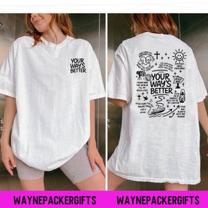 Puede incluir: Camiseta blanca con el texto "YOUR WAY'S BETTER" en la parte delantera y un diseño gráfico en la parte trasera. El diseño trasero incluye texto e ilustraciones de una cruz, un sol, un barco y otros símbolos. La marca "WAYNEPACKERGIFTS" está en la parte inferior.