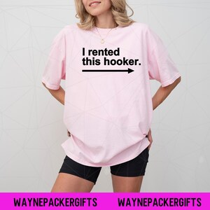 Puede incluir: Camiseta rosa claro con el texto negro "I rented this hooker" y una flecha hacia la derecha. La camiseta se combina con pantalones cortos negros. La parte inferior de la imagen muestra "WAYNEPACKERGIFTS" en blanco sobre un fondo rosa.