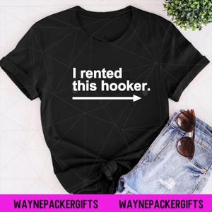 Puede incluir: Camiseta negra con el texto blanco "I rented this hooker" y una flecha que apunta hacia la derecha. La camiseta se combina con pantalones cortos vaqueros y gafas de sol.