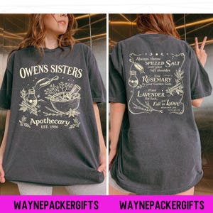 May include: A charcoal gray t-shirt featuring a cream-colored graphic print. The front displays the text "OWENS SISTERS APOTHECARY EST. 1986" with illustrations of herbs and a mortar and pestle. The back features text about herbs and love.