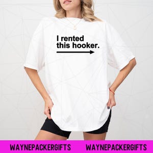 Puede incluir: Camiseta blanca con el texto "I rented this hooker" en negro, con una flecha que apunta hacia la derecha. La camiseta la lleva una persona. La parte inferior de la imagen tiene el texto "WAYNEPACKERGIFTS" en blanco sobre un fondo rosa.