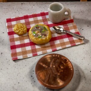 May include: A miniature soup bowl with a red and white checkered napkin, a small bread roll, a white ceramic mug, and a silver spoon. A penny is in the foreground.