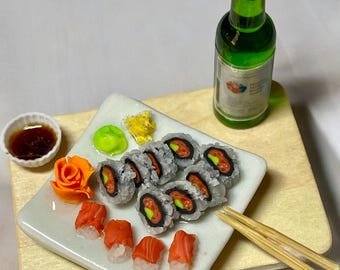 Puede incluir: Un plato blanco con una porción de rollos de sushi, nigiri de salmón y un acompañamiento de jengibre encurtido. Los rollos de sushi están cubiertos con aguacate y carne de cangrejo de imitación. El plato está sobre una tabla de cortar de madera con una pequeña botella de soju.