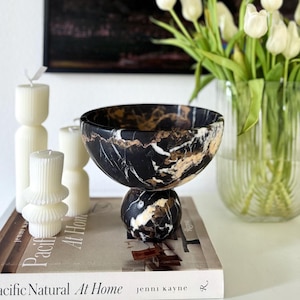 May include: A black and gold marble pedestal bowl sits atop a book titled Pacific Natural At Home by Jenni Kayne. Beside it are three white ribbed pillar candles of varying heights and a clear ribbed glass vase filled with white tulips.
