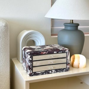 May include: A rectangular jewellery box with a dark purple and white marbled stone exterior featuring three horizontal drawers with silver-toned metal fronts and small matching knobs, positioned next to a white arched object and a grey table lamp.