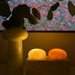 Puede incluir: Dos lámparas de piedra ovaladas y brillantes, en tonos amarillos y naranjas, descansan sobre una base rectangular de piedra. A la izquierda, un jarrón alto y claro con una ramita de eucalipto. Un cuadro colorido en el fondo.