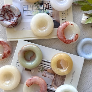 May include: A collection of decorative, donut-shaped stone coasters in various colors, including white, yellow, pink, and green. Some coasters have veining patterns. The coasters are arranged on a book.