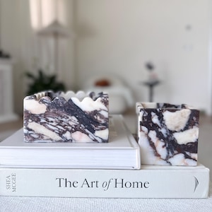 Puede incluir: Dos recipientes de mármol con un patrón veteado blanco y morado oscuro se asientan sobre una pila de libros. El libro de abajo tiene el título "The Art of Home" en texto negro. Los recipientes tienen forma cuadrada.